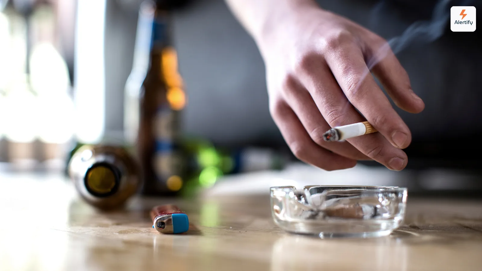 Gast hält eine brennende Zigarette über einen Glasaschenbecher neben leeren Flaschen auf einem Tisch