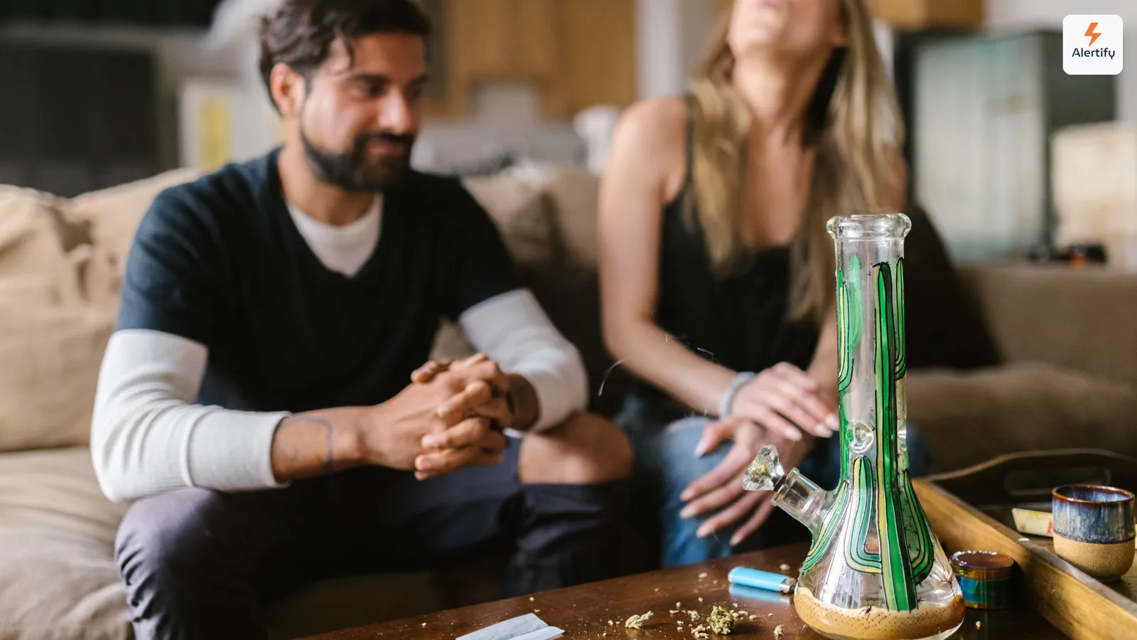 Zwei Gäste sitzen auf einem Sofa im Wohnzimmer einer Ferienwohnung, auf dem Couchtisch vor ihnen eine Glasbong, ein Feuerzeug und ein Grinder.